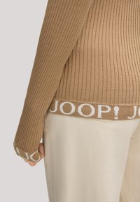 Rippbrauner Pullover mit weißem "JOOP!"-Logo am Saum, ausgestattet mit langen Ärmeln und einer strukturierten Optik. Kombiniert mit hellen Hosen.