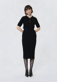 Γυναίκα που φοράει ένα μαύρο φόρεμα κοντό μανίκι με ζώνη, διαφανείς καλσόν με σχέδιο καρδιές, μαύρες μυτερές γόβες, κολιέ με μαργαρίτες και μαύρη κορδέλα στο κεφάλι.