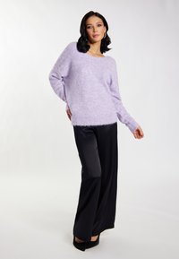 Lila, flauschiger Pullover mit weitem Ausschnitt und geripptem Saum, kombiniert mit glänzenden schwarzen weiten Hosen und schwarzen Absätzen.