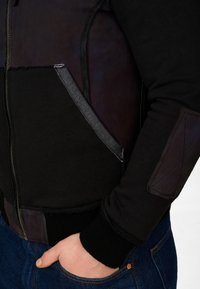 Svart jacka som kombinerar läder och sweatshirting, med zip-förslutning, sidofickor och texturerade stickade manschetter. Mörka och kontrasterande accenter.