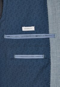 Marineblauwe stof met een subtiel geometrisch patroon, met een label waarop "MODERN FIT" staat en twee horizontale zakken met contrasterende blauwe randen.