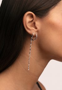 Zilveren oorring in de vorm van een ring met een hangende ketting die afwisselend geometrische vormen en heldere stenen bevat, getoond tegen een huidkleurige achtergrond.