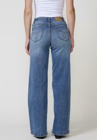 Jeans de mezclilla azul de pierna ancha con un lavado claro. Presentan bolsillos traseros con costuras decorativas y un parche de cuero marrón en la cintura.