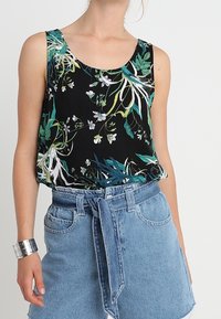 Top senza maniche floreale con sfondo nero, caratterizzato da motivi vegetali verdi e bianchi; indossato con una gonna di jeans a vita alta e un bracciale argento.