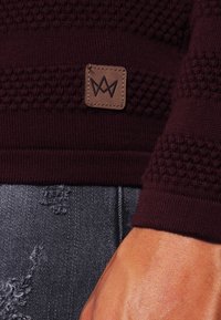Bordowy sweter dzianinowy z teksturowanym wzorem i gładką dolną częścią; posiada brązową skórzaną metkę z logo; zestawiony z postrzępionymi szarymi dżinsami.