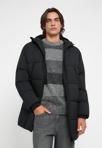 Giacca a vento nera con cappuccio, caratterizzata da un design trapuntato, chiusura con zip e tasche laterali, indossata sopra un maglione grigio a righe.