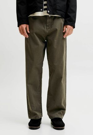 Adulte portant un pantalon ample vert olive, des chaussures noires, une chemise rayée et une veste noire, debout sur un fond clair uni.