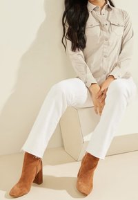 Camisa de botones color beige claro con dos bolsillos en el pecho, combinada con jeans blancos de pierna recta y botas de tobillo de ante marrón con tacones tipo bloque.