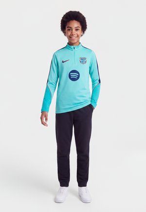 Enfant souriant debout portant un haut de sport turquoise avec les logos de Barcelone et Spotify, pantalon noir et baskets blanches sur fond blanc.