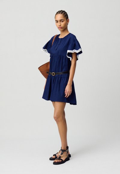 Un vestido azul marino con un diseño de mangas acampanadas y ribete blanco, ceñido en la cintura con un cinturón negro. La modelo lleva sandalias negras y lleva un bolso marrón.
