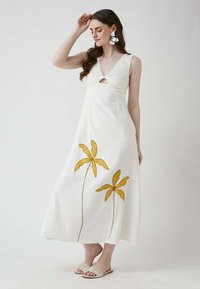 Weißes Leinenkleid mit V-Ausschnitt und einem Cut-out an der Vorderseite. Verziert mit gelben, gestickten Blumen am Saum. Kombiniert mit cremfarbenen Schlüpfsandalen.