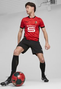 Camiseta de fútbol roja con un diseño de telaraña, detalles en negro y logo blanco; pantalones cortos y calcetines negros; el jugador está de pie con un balón rojo y verde.