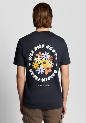 Persoon met kort bruin haar, gekleed in een donker T-shirt met kleurrijke bloemmotieven en de tekst "Lyle and Scott Garden Fresh Since 1874" op de achterkant.