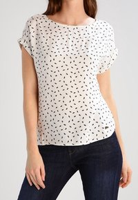Blusa crema con stampa nera a foglie, maniche corte arrotolate, scollatura rotonda e orlo curvo. Abbinata a jeans scuri.