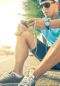 Un uomo casual seduto su uno skateboard, indossa una maglietta blu e pantaloni corti neri, mentre usa un telefono con auricolari. Scarpe da ginnastica grigie con suola bianca.