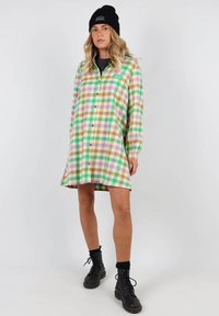 Robe chemise à carreaux en vert, violet, brun et blanc. Manches longues, ourlet arrondi, portée avec des bottes de combat noires et un bonnet.