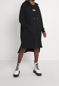 Schwarzes oversized Kapuzenkleid mit einer Vordertasche, seitlichen Schlitzen und einem Logo auf der Brust. Kombiniert mit weißen Schnür-Stiefeln.