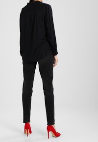 Black long-sleeve blouse with a rounded hem, paired with fitted black jeans and red high-heeled shoes. Smooth fabric with subtle draping.