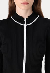 Melna ribota auduma blūze ar augstu apkakli, kas izceļas ar baltu rāvējslēdzēju un akcentu apmalēm apkakles malā. Gluda tekstūra un piegulošs dizains.