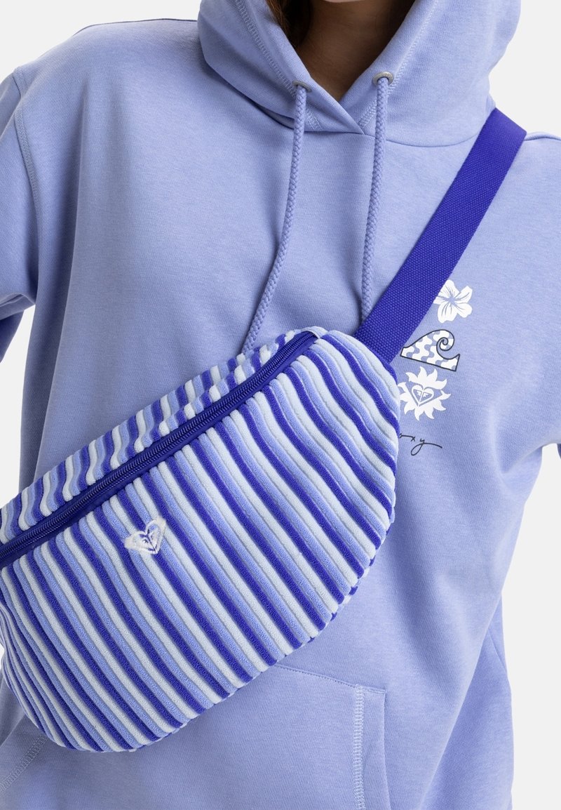 Personne portant un sweat à capuche lilas avec des graphiques floraux, portant un sac bandoulière rayé bleu et blanc avec une fermeture éclair et un logo brodé.