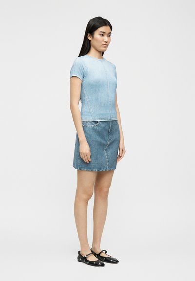 Γυναίκα που στέκεται φορώντας εφαρμοστό ανοιχτό μπλε κοντομάνικο τοπ, φούστα από τζιν και μαύρα επίπεδα παπούτσια με ασημένιες τρύπες σε γκρι φόντο.