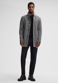 Cappotto in misto lana grigio con collo alto, bottoni frontali e taglio dritto. Il modello indossa un maglione a maglia nera e pantaloni neri a sigaretta.
