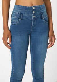 Jeans de mezclilla azul de cintura alta con cierre de doble botón, bolsillos frontales y una textura suave con un ligero desvanecimiento en los muslos.