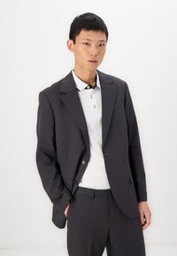 Grijze op maat gemaakte blazer met een gestructureerde pasvorm, voorzien van een geknipt revers, twee knopen, en gecombineerd met een wit poloshirt met een zwarte kraag.