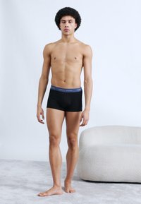 Μαύρα ανδρικά εσώρουχα boxer με ανοιχτό γαλάζιο ελαστικό μέση, με λογότυπο, φορεμένα από άνδρα μοντέλο που στέκεται σε απαλό γκρι χαλί.