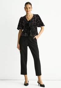 Black sequined top with puff sleeves and a V-neck, featuring a front bow. Paired with fitted black trousers and black heeled shoes.