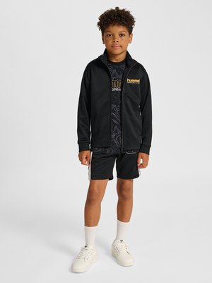 Dreng iført sort lynlåstrøje, mønstret sort t-shirt, sorte shorts med hvide striber, hvide sokker og hvide sneakers står mod en ensfarvet baggrund.