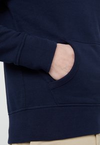 Pravá ruka osoby v přední kapse tmavě modré mikiny, s béžovými kalhotami viditelnými pod mikinou.