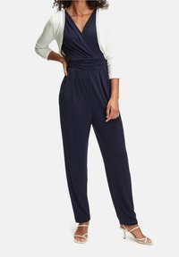 Mono de marina con un cuerpo de diseño envolvente, cintura ajustada y pantalones anchos. Combinado con un cárdigan de color claro y tacones con tiras.