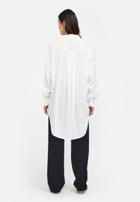 Blusa bianca a maniche lunghe realizzata in tessuto leggero, con orlo arcuato, spalla posteriore e vestibilità rilassata con polsini bottonati.