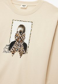 Beżowy sweatshirt z grafiką przedstawiającą widok z tyłu, featuring związana wstążka w leoparda, oprawiona czarną i złotą linią, z tekstem poniżej.