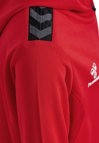 Szczegół ramienia czerwonej sportowej koszulki z czarno-białym wzorem chevron na teksturowanej tkaninie, pokazujący część białego logo na klatce piersiowej.