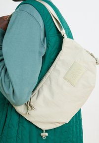 Persona que lleva un chaleco acolchado en color teal y un sudadera azul-verde, porta una bolsa de hombro beige de The North Face con correa ajustable.