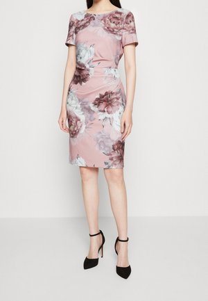 Kobieta w dopasowanej różowej sukience do kolan z dużymi białymi i bordowymi kwiatowymi wzorami oraz czarnych wysokich obcasach z paskiem wokół kostki.