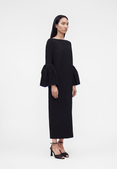 Γυναίκα που στέκεται φορώντας μακρύ μαύρο φόρεμα με φαρδιά μανίκια καμπάνα και μαύρα πέδιλα με τακούνια μπροστά σε απλό ανοιχτό φόντο.