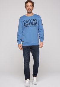 Felpa blu a girocollo con testo nero "2501 NY UK CAMP DAVID" e dettagli aggiuntivi, abbinata a jeans scuri e sneakers bianche.