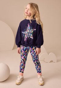 Navy sweatshirt with colorful star graphic, paired with multi-colored floral leggings. White sneakers with beige accents and pink socks.