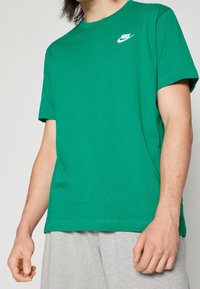 Osoba v zeleném tričku Nike a svetle šedých teplákách, s uvolněnýma rukama podél těla, na jednobarevném pozadí.