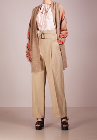 Pantalons larges beiges avec une taille ceinturée, associés à un blouse à volants rose clair et un cardigan rayé marron et orange. Sandales à plateforme noires.