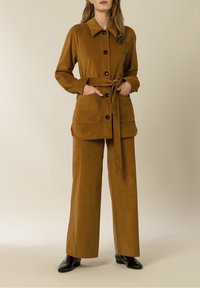 Ruskea corduroy-asuste, johon kuuluu pitkähihainen nappikauluspusero, jossa on kaulus, solmittava vyö ja kaksi etutaskua, yhdistettynä leveälahkeisiin housuihin.