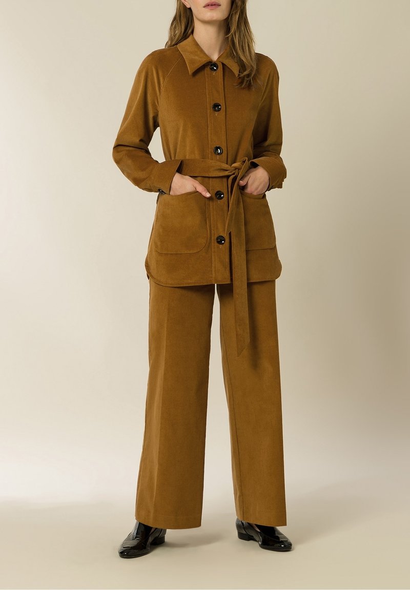 Ruskea corduroy-asuste, johon kuuluu pitkähihainen nappikauluspusero, jossa on kaulus, solmittava vyö ja kaksi etutaskua, yhdistettynä leveälahkeisiin housuihin.