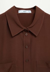 Blouse marron avec un col, fermeture à boutons sur le devant, et deux poches poitrine. Fabriquée en tissu lisse avec une texture subtile.