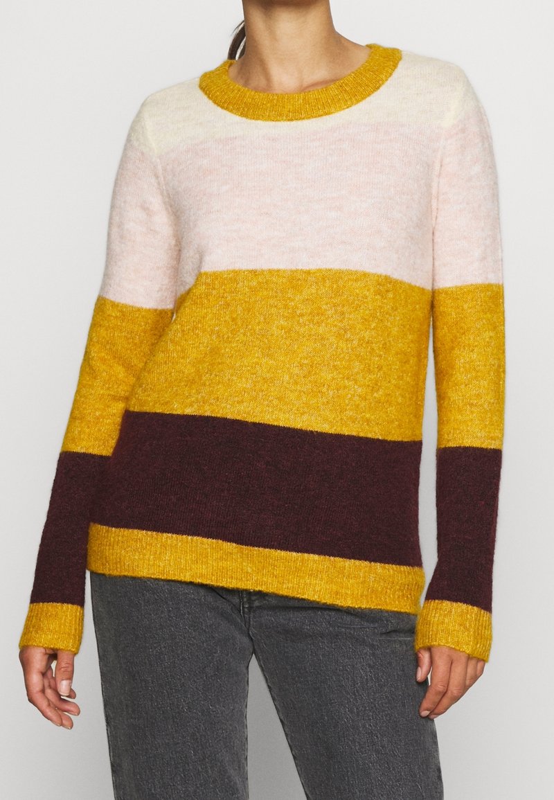 Femme portant un pull en tricot à manches longues avec des rayures horizontales crème, jaune moutarde et brun foncé, associé à un pantalon gris foncé.