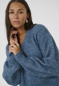 Pullover a maglia blu con texture, scollo a V, maniche larghe e polsini a coste, indossato da una persona con capelli di lunghezza media.
