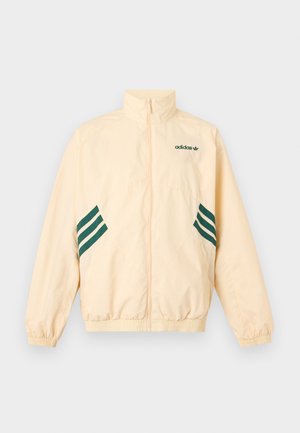 Lichtbeige Adidas trainingsjack met hoge kraag, volledige rits aan de voorkant, elastische boorden en zoom, en donkergroene Adidas-logo en strepen aan de zijkanten.