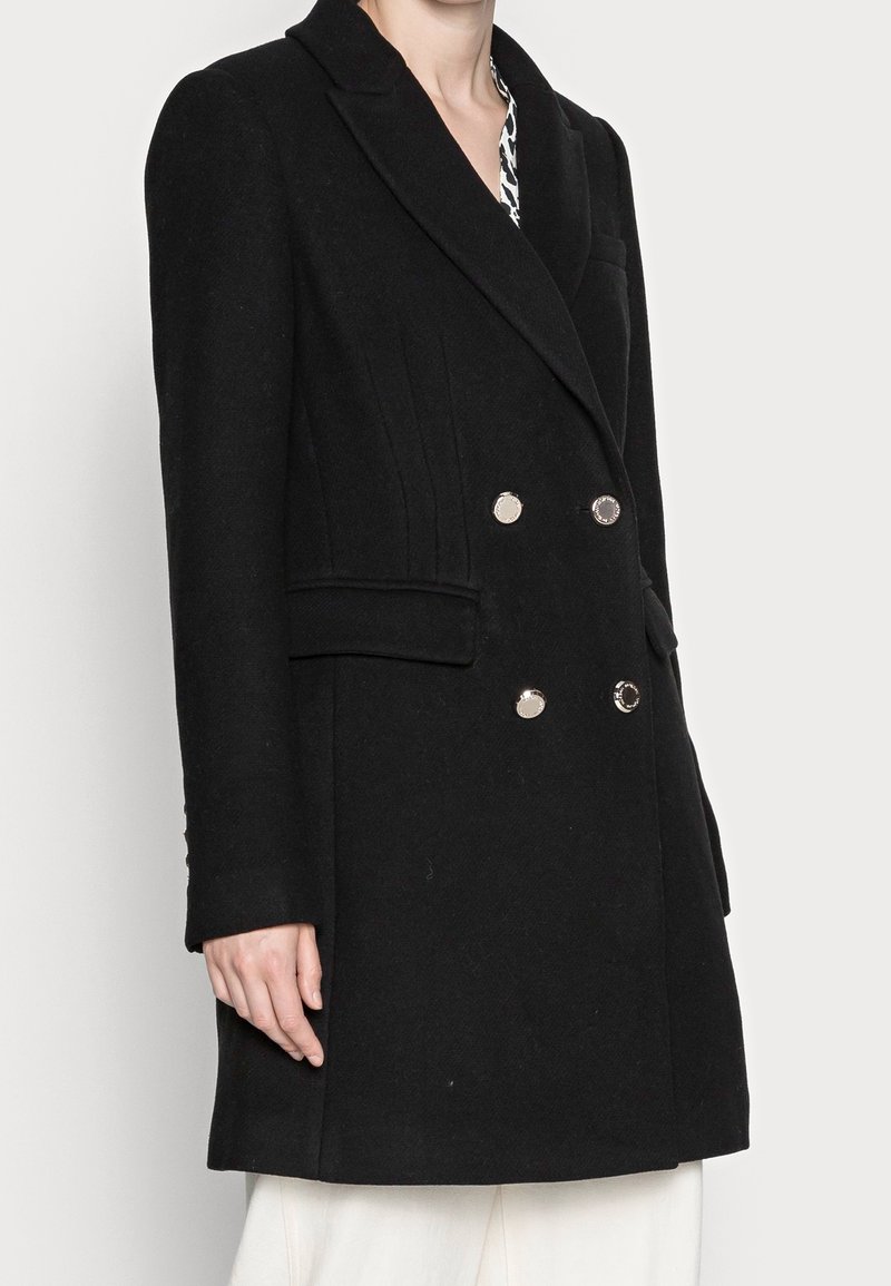 Manteau noir croisé à double boutonnage avec un col cranté, des boutons argentés et des poches latérales. En laine, il présente des coutures verticales et des manches longues.